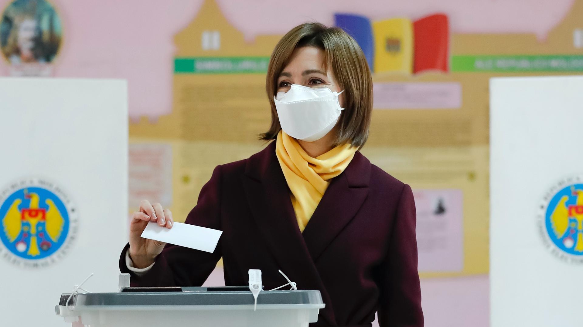 A woman wearing a suit with yellow shirt casts a ballot while wearing a mask