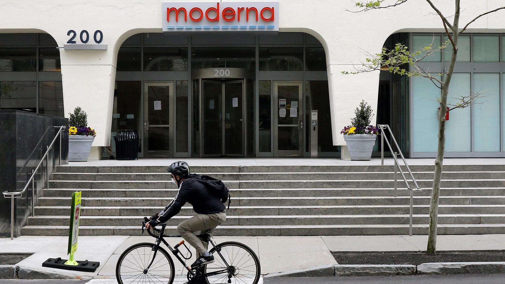 A bicyclist wearing a dark jacket and helmet is shown riding past the outside of drugmaker Moderna.