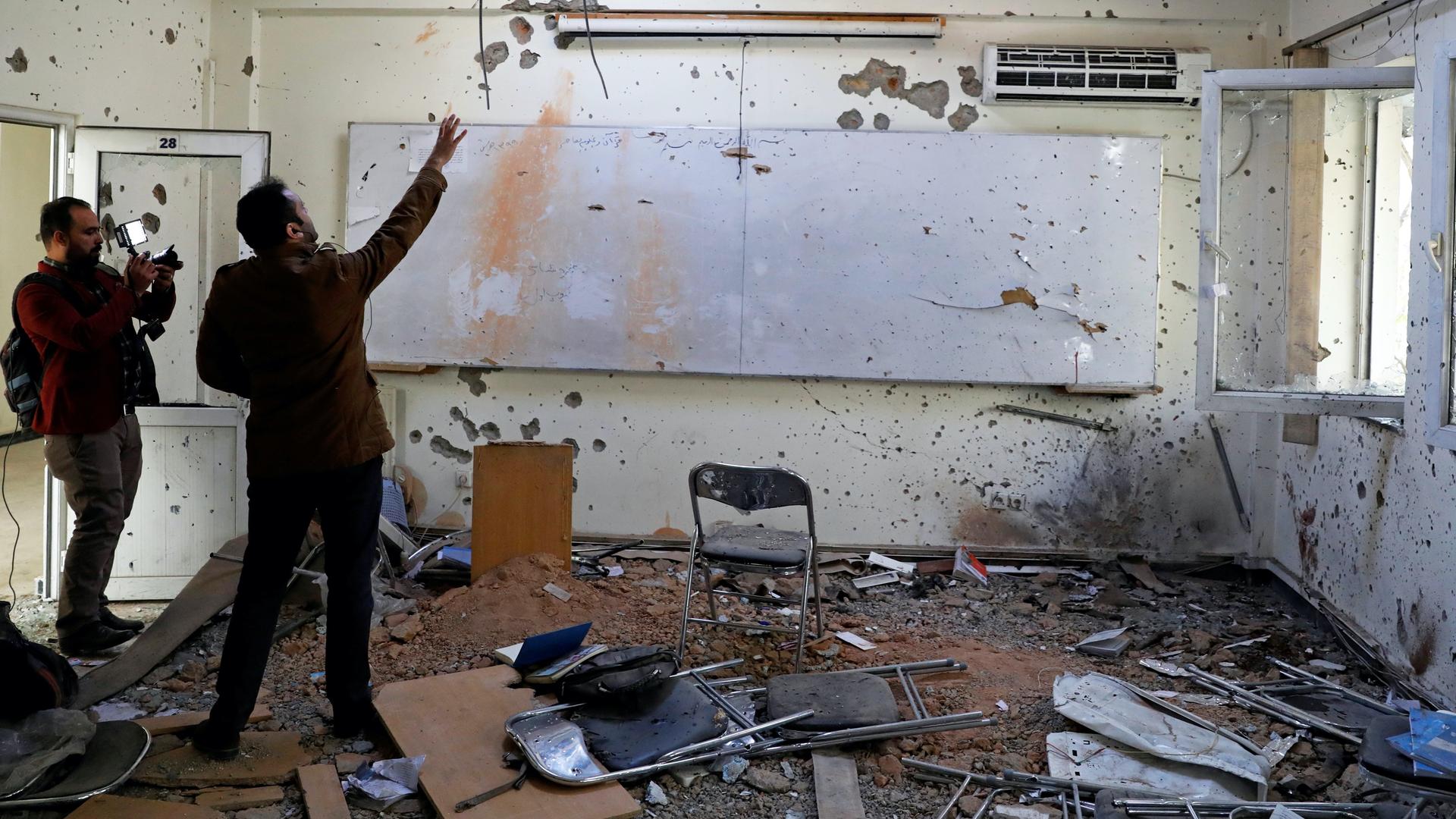 Two journalists look at a bombed out classroom with chair strewn and walls shot with bullets. 