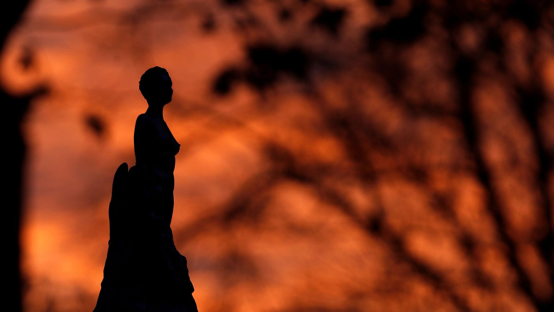 The dark silhouetted of a statue is shown with the orange and red colors of a sunset in the distance.