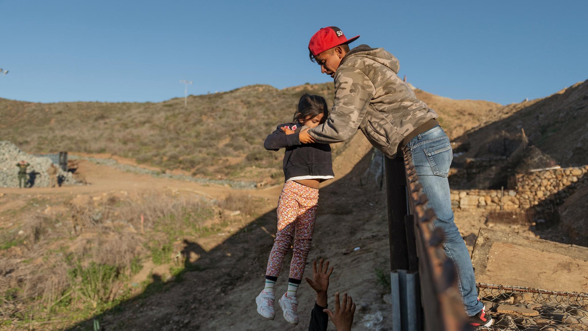 A child is passed over a border wall to the arms of a man wearing jeans, a red cap and tan sweatshirt. 