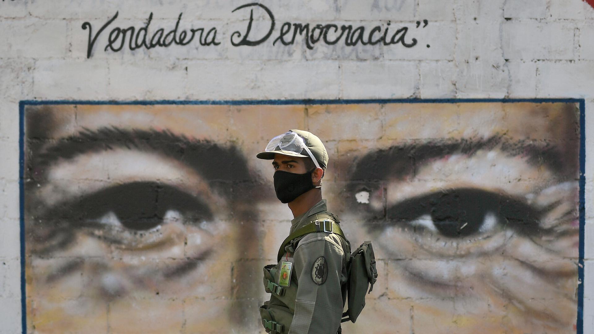A soldier is shown wearing a face mask and a military uniform while standing in front of a wall painted with the eyes of the late President Hugo Chávez.