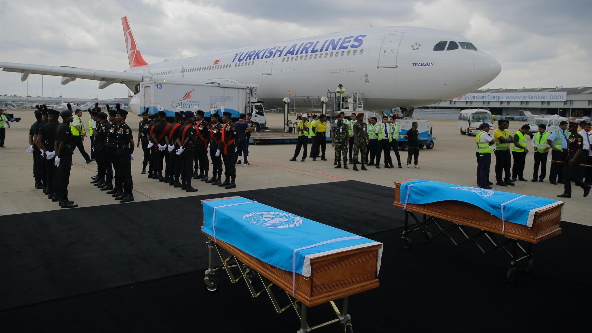 Two caskets covered with UN flags on display near a Turkish airlines plane and military personel standing nearby. 