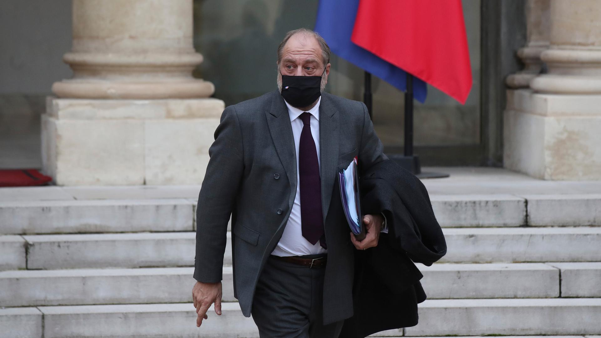 French Justice Minister Eric Dupond-Moretti is shown wearing a dark-colored face mask and gray suit while carrying a folder.