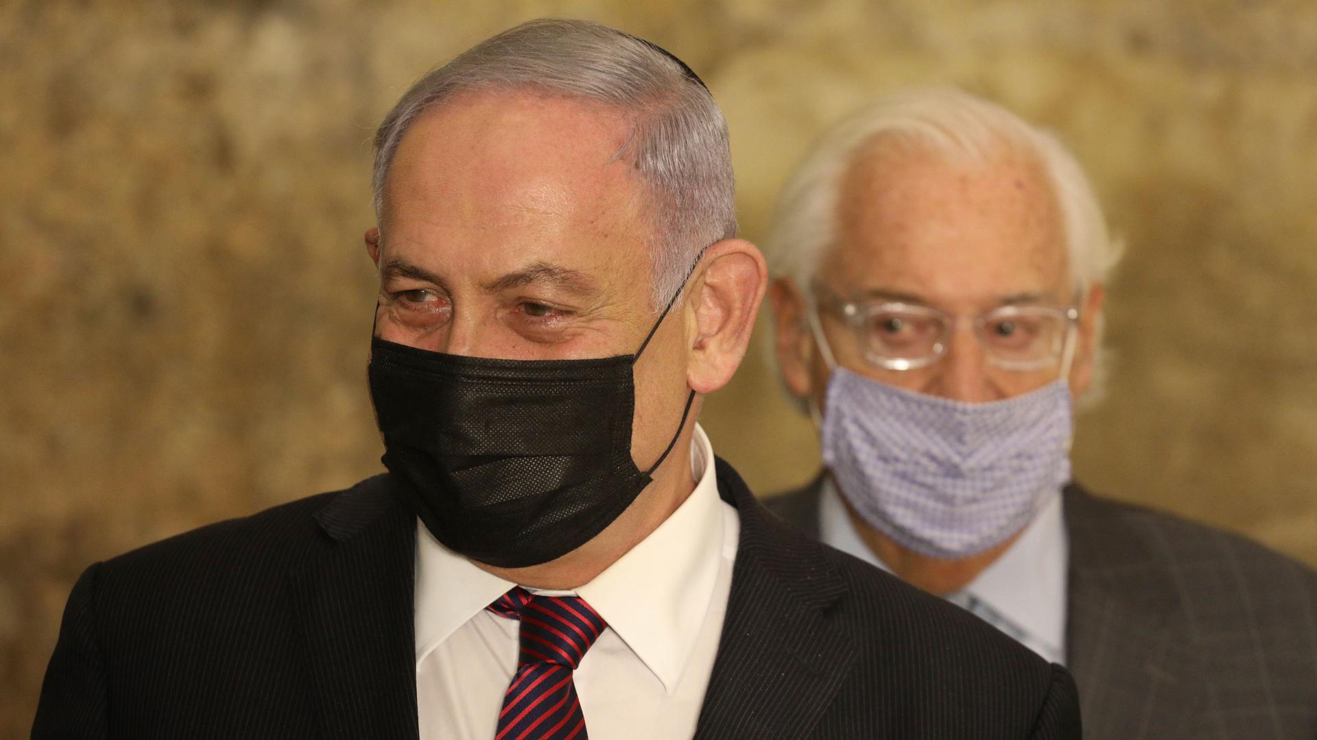 Israeli Prime Minister Benjamin Netanyahu is shown wearing a face mask and suit with US Ambassador to Israel David Friedman standing behind him.