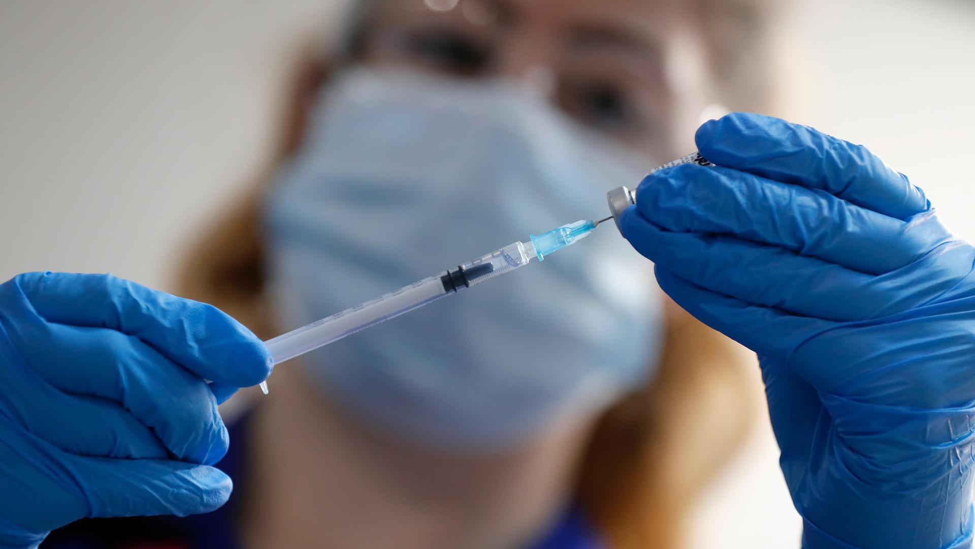 A close-up photograph show a syringe being held by two hands wearing blue medical gloves and the face of a nurse in soft focus in the background.