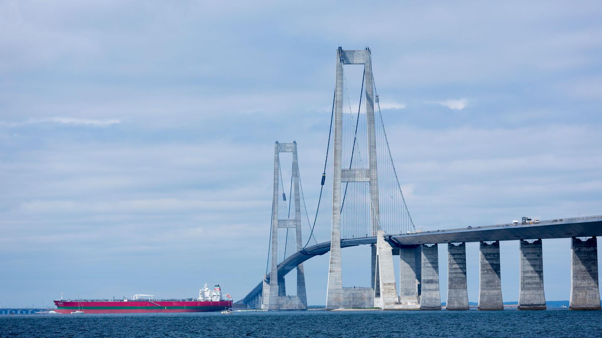 A large red oil tanker is shown in the distance passing underneath two tall bridge columns.