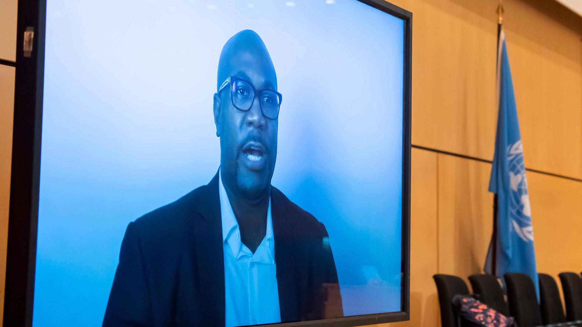 A Black American man wears glasses and a blue suit jacket and speaks via video with a blue background 