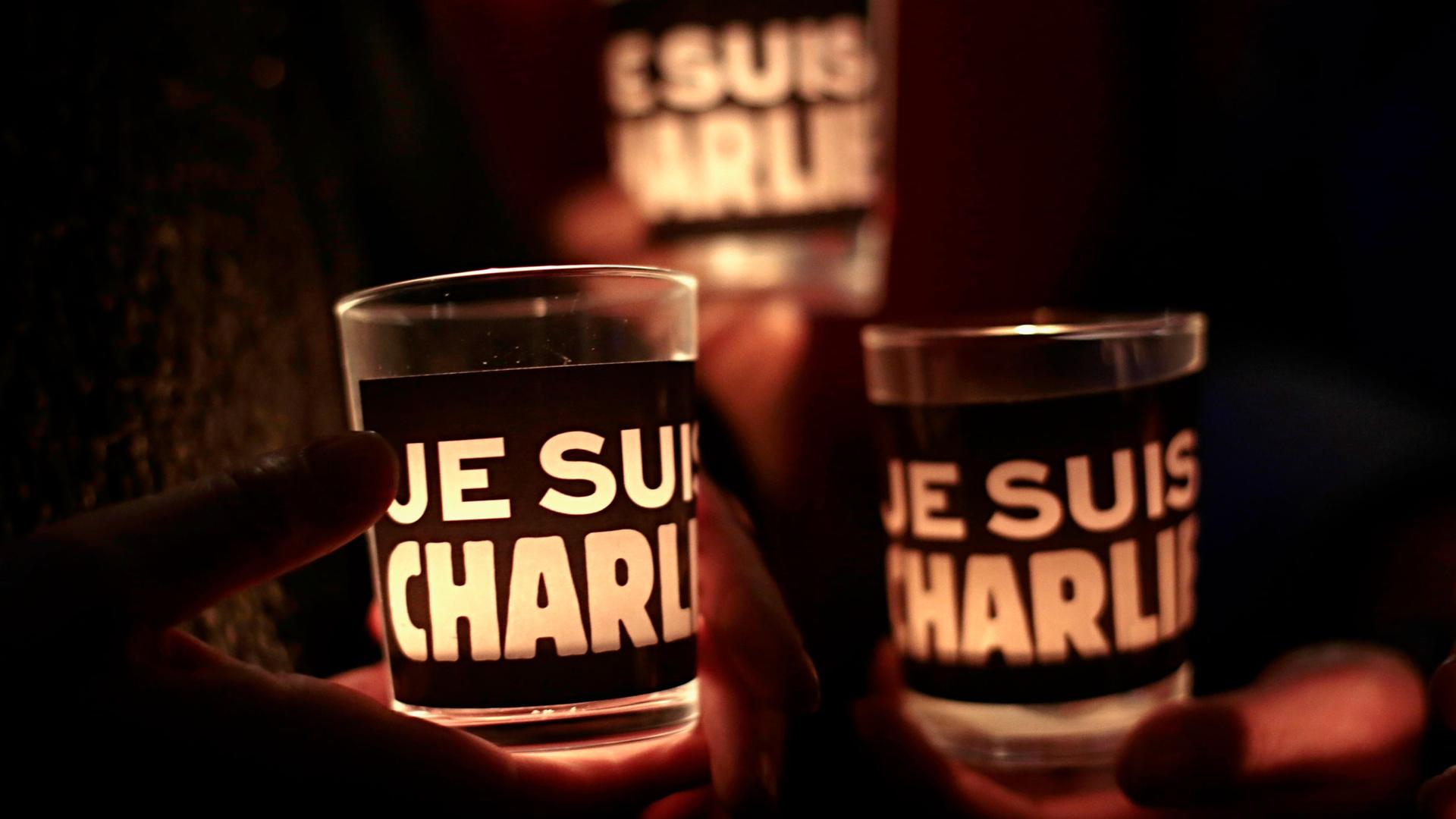 In a photograph taken in the dark, three glasses with candles glowing inside are shown with the phrase 'Je Suis Charlie' printed on it.