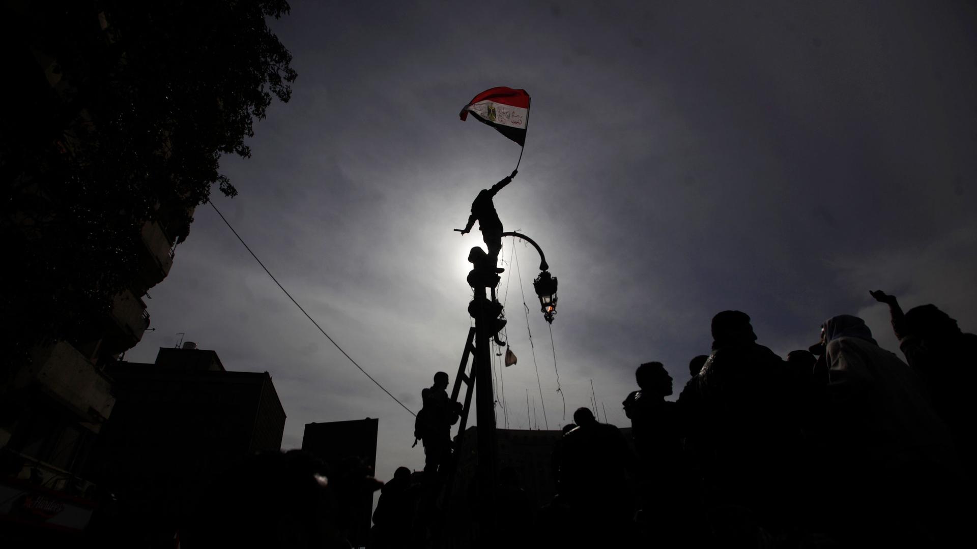 A protester is shown standing on top of a street light and holding the Egyptian flag while blocking the sun and creating a shadow.
