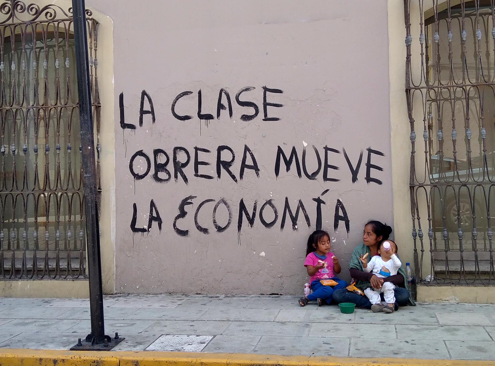 A woman with children asks passersby in downtown Oaxaca for spare change next to a wall painted with a slogan that reads 