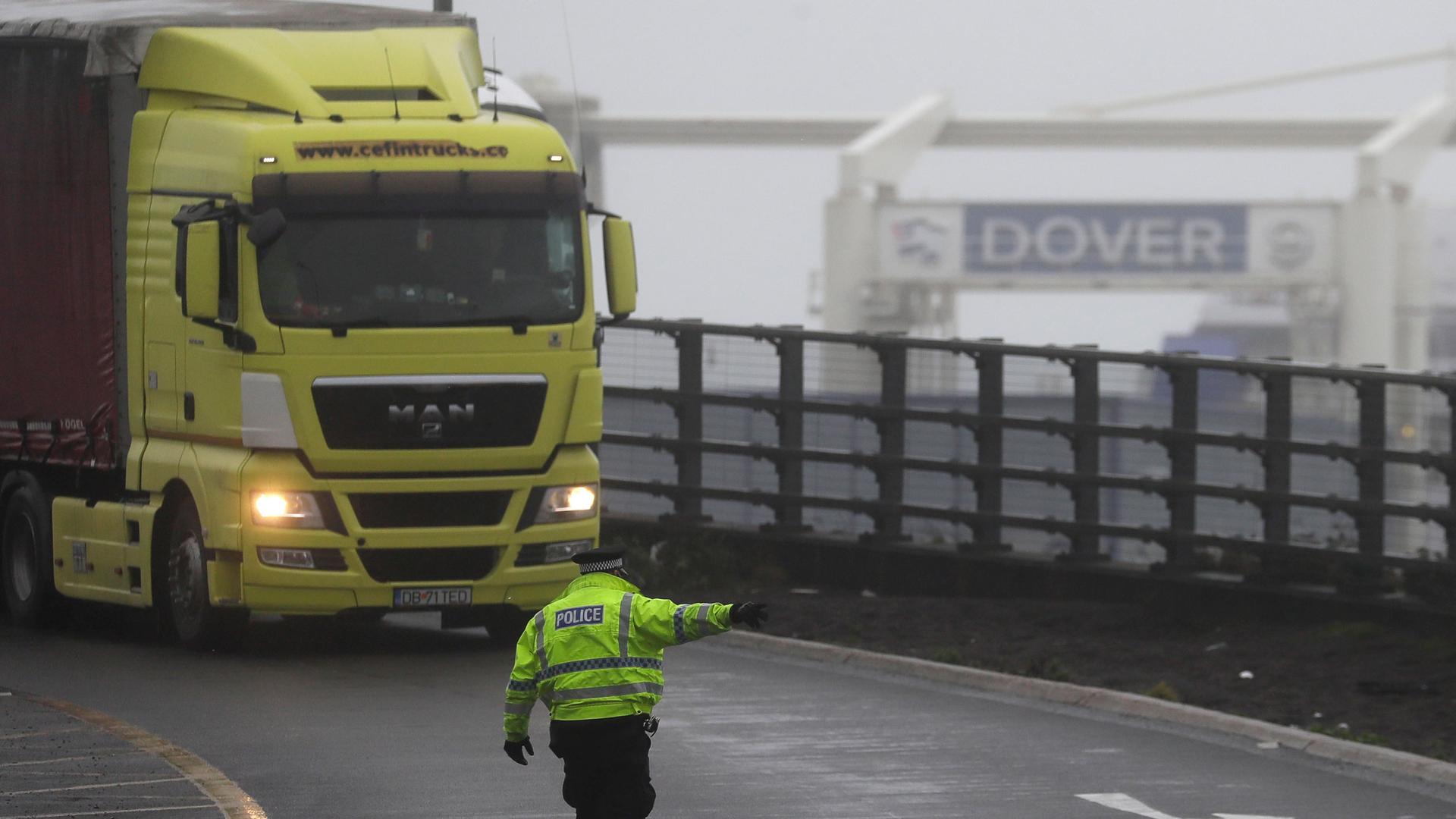 A yellow semi-truck is shown being directed by a police officer wearing a reflective jacket with a sign for Dover in the background.