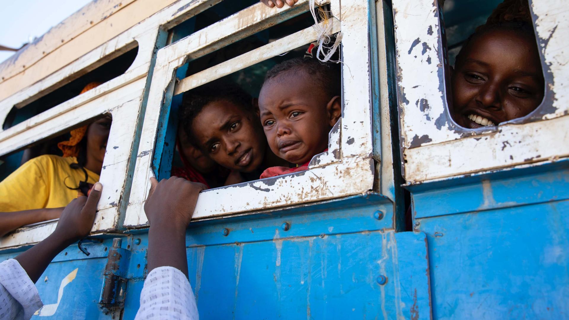 Three Ethiopian travelers sit inside a blue bus with white-bordered windows, saying goodbye to someone whose arms rest on the window frame. 