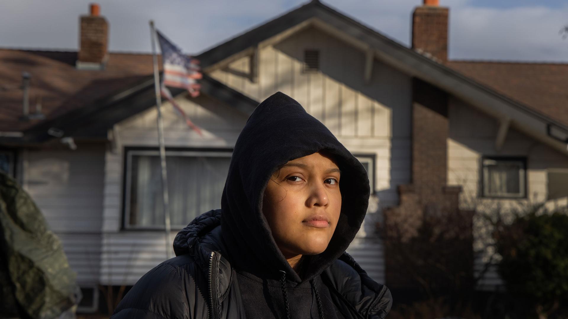A woman is shown wearing a dark hooded sweatshirt and jacket with a home in the distance flying a tattered US flag.