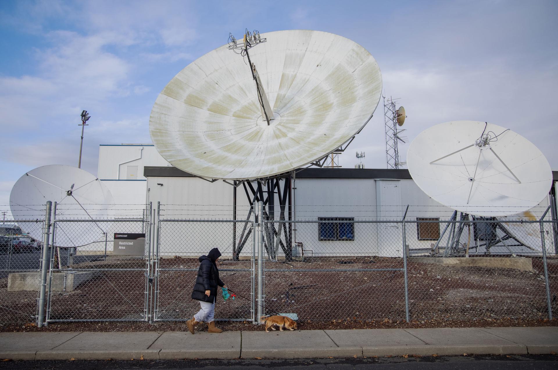 A woman is shown walking a small brown dog with a chain link fence and large satellite dishes in the distance.