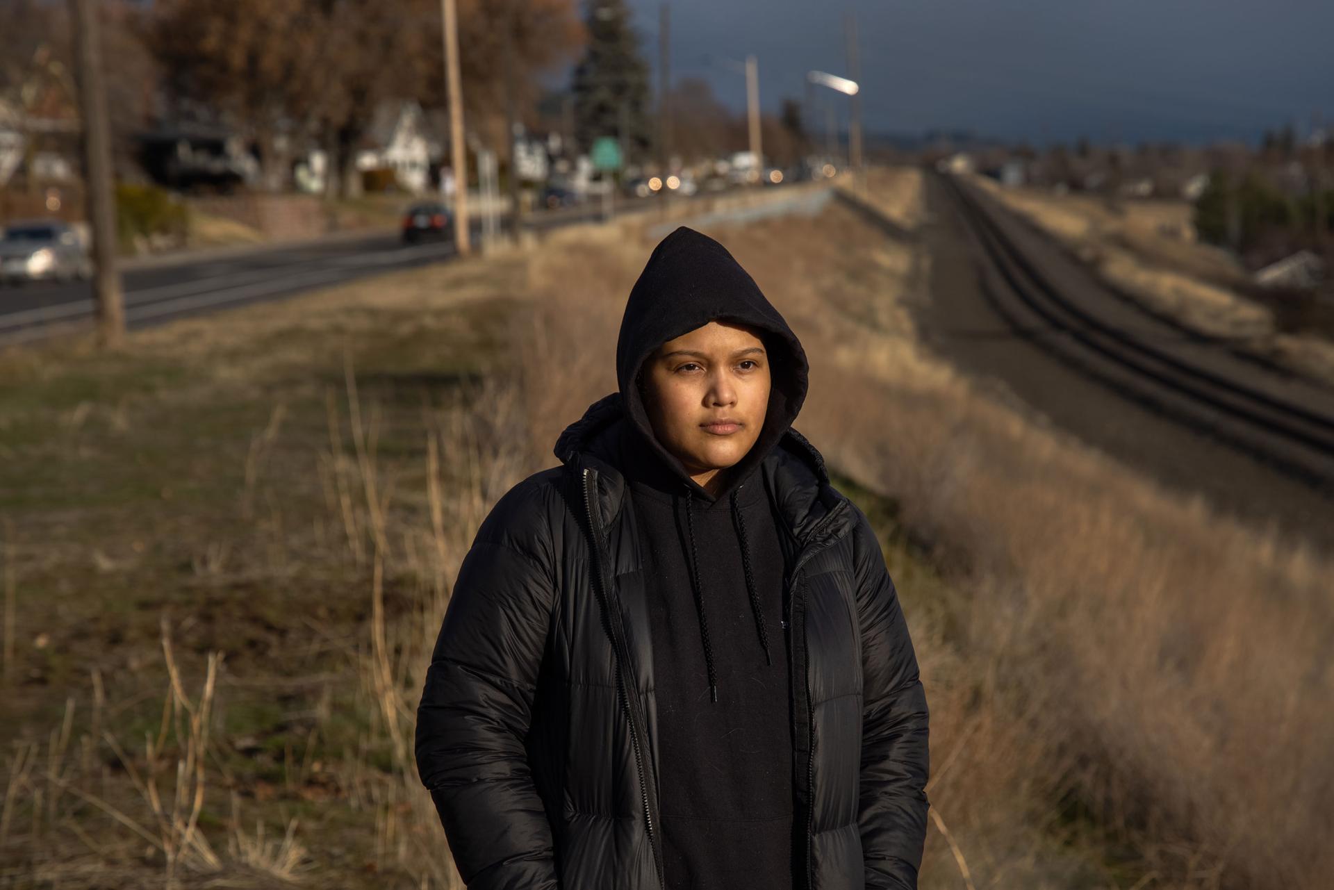 A woman is shown wearing a dark hooded sweatshirt and jacket while standing near a set of railroad tracks.