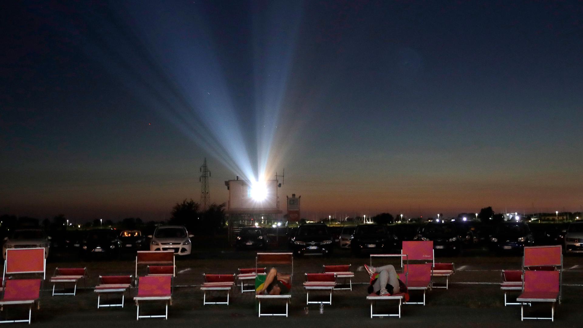 Several rows of outdoor seating are shown with cars in the distance and a movie project shining bright in the distance.