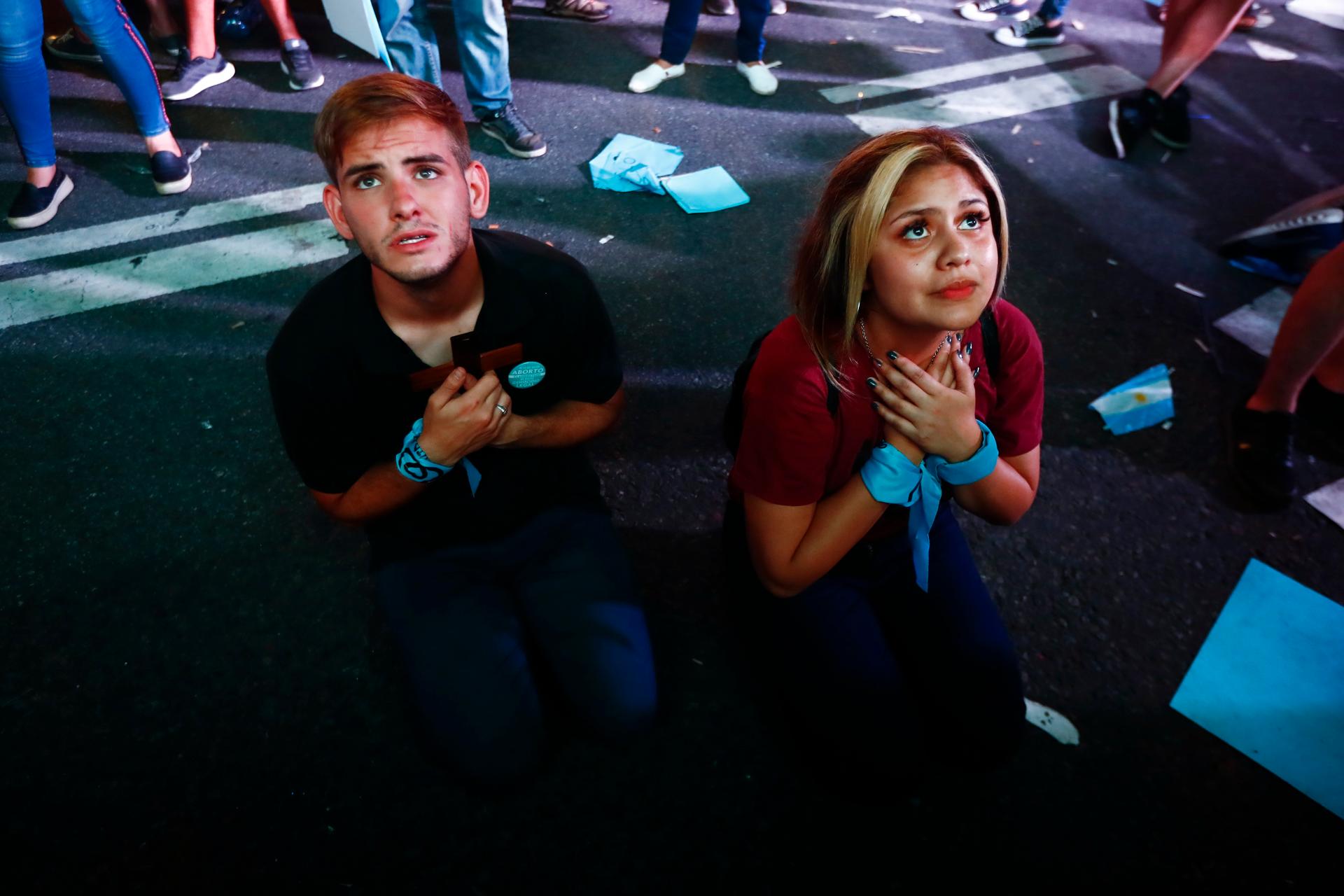 A young man and a young woman, wearing a blue scarf around their wrist, kneel down to pray on the street.