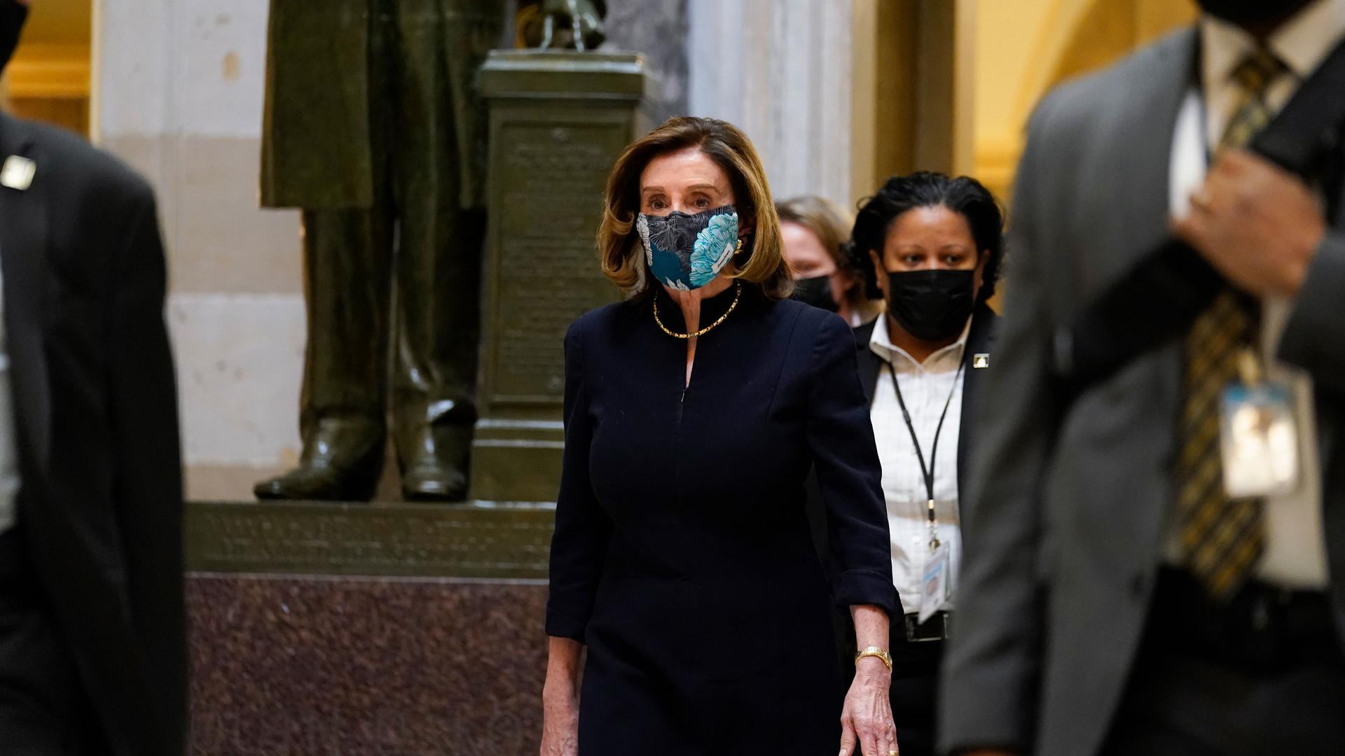 Speaker of the House Nancy Pelosi is shown wearing a dark suit and a face mask while walking in the halls of Capitol Hill.