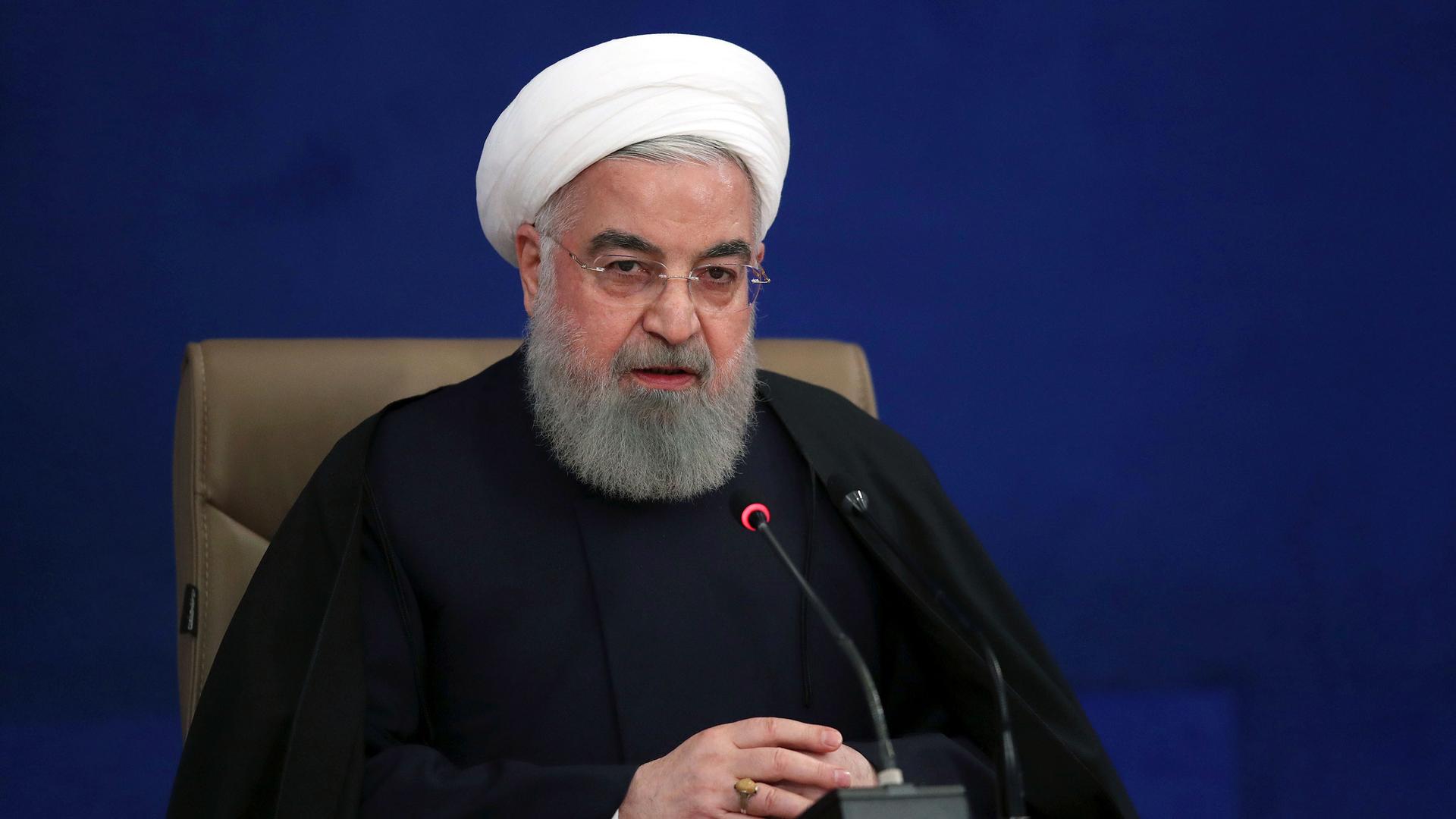 Iranian President Hassan Rouhani is shown wearing a traditional white turban while sitting behind a microphone.
