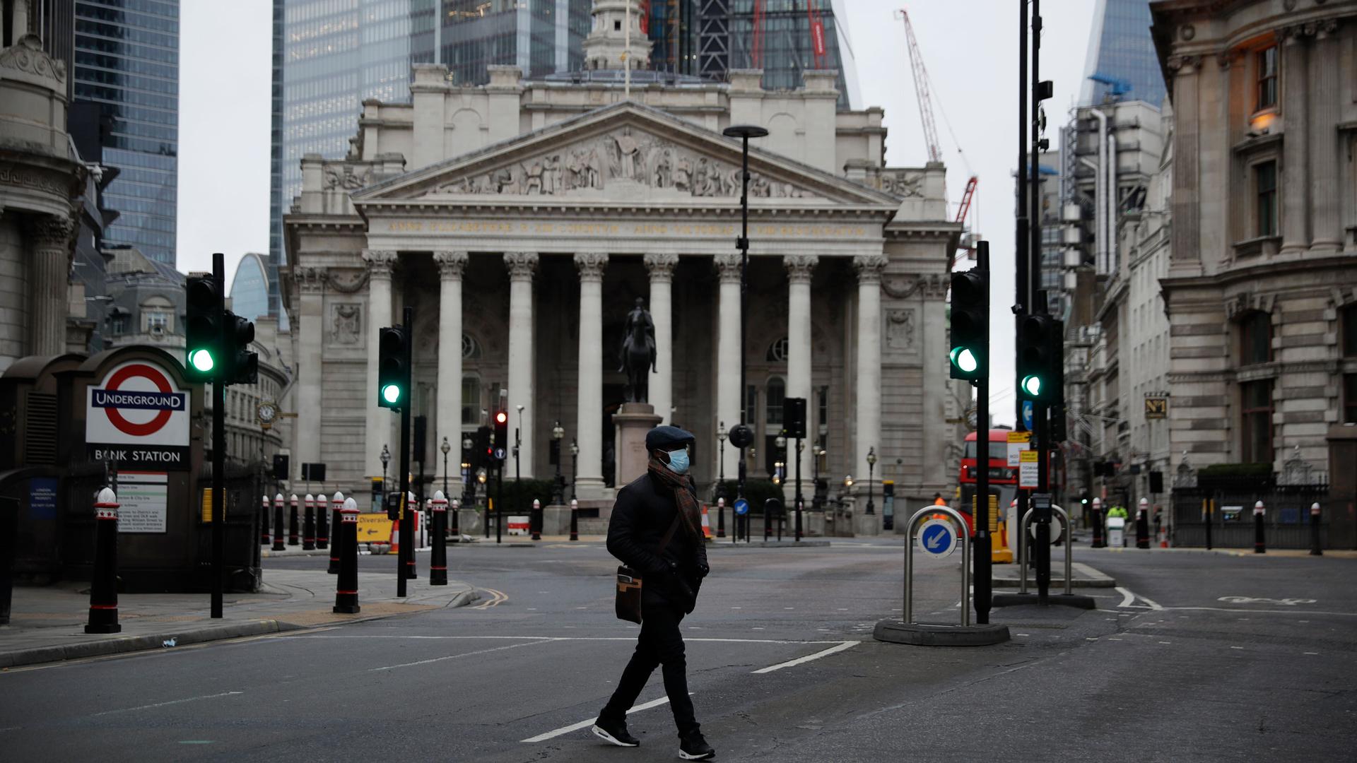 A man is shown with his hands in his coat pockets and wearing a face mask crossing a the street with England's Royal Exchange is in the distance.