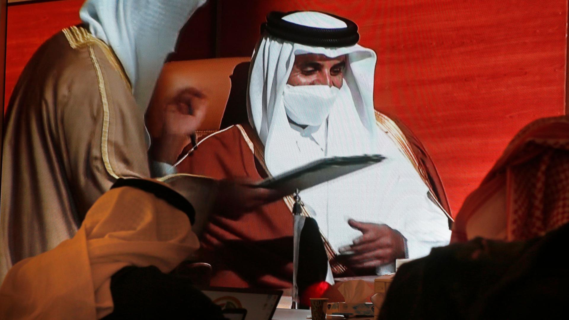 Two people are shown from behind wearing head coverings and looking up at a screen showing with Qatar's Emir Sheikh Tamim bin Hamad Al Thani.