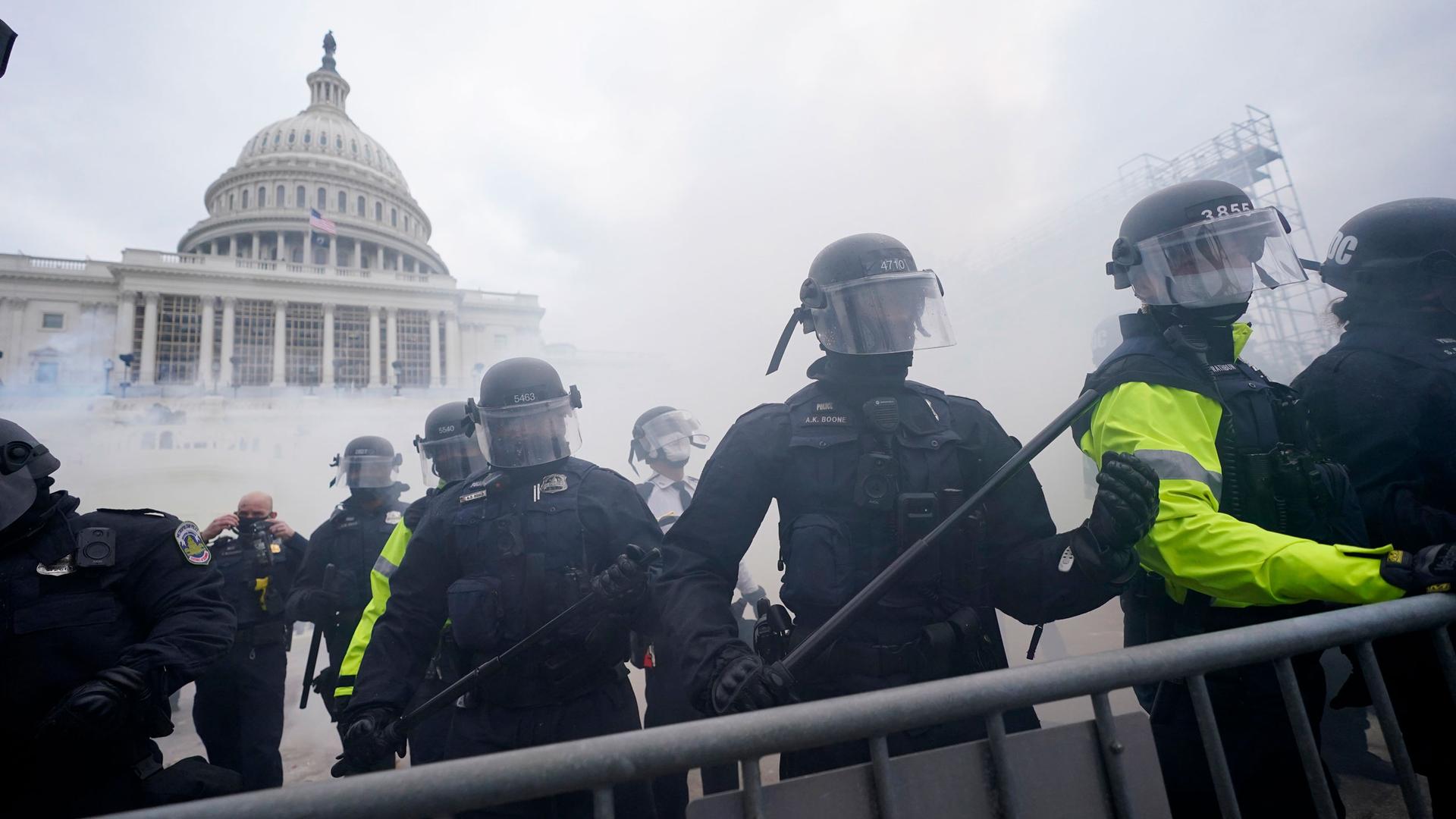 The US Capitol building is shown with smoke and riot police standing behind a baracade.