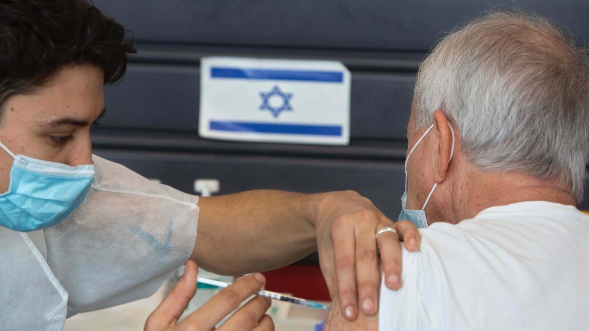 A health worker gives a shot in the arm to a person with silver hair in front of a blue and white Israeli flag. 