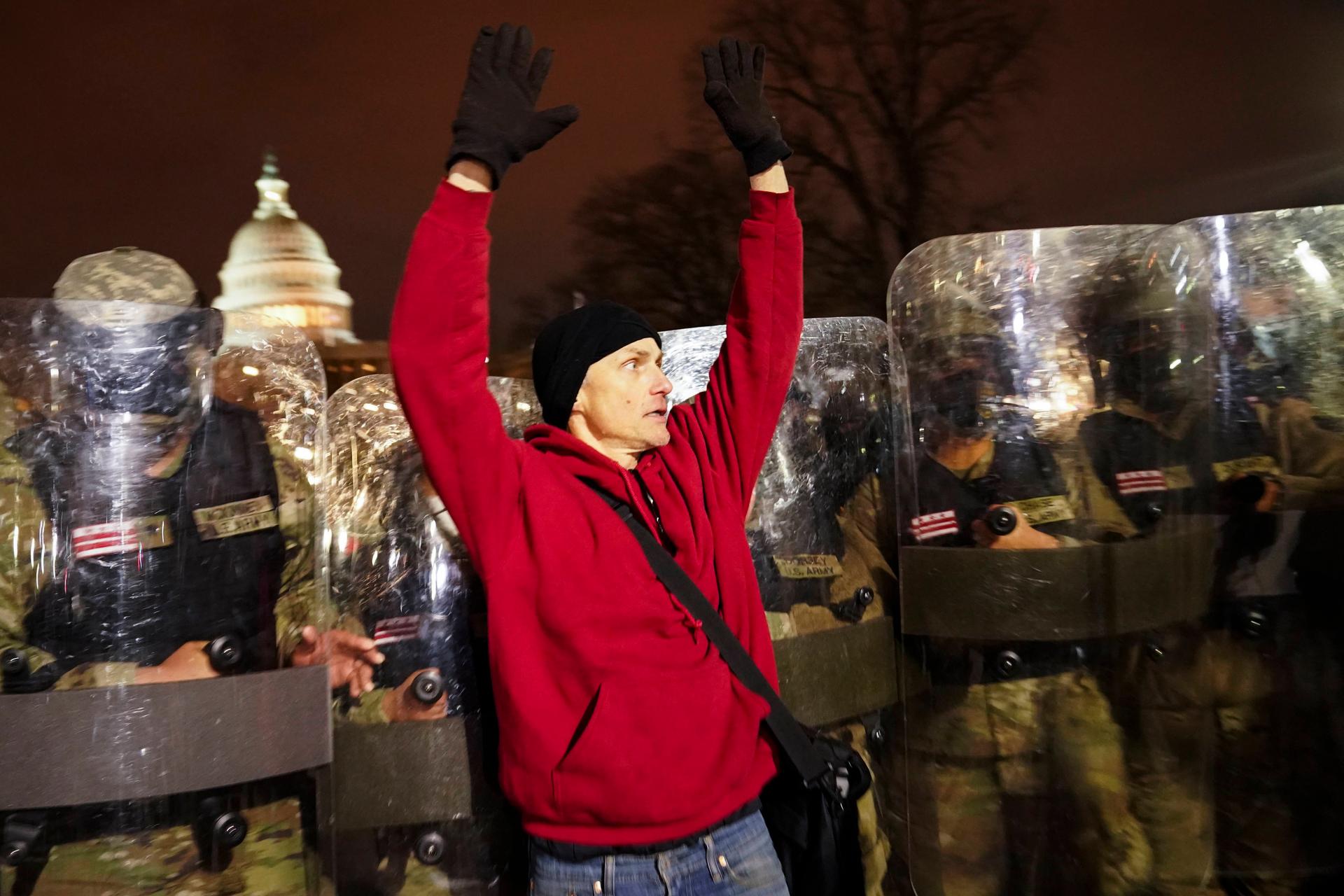 National guard appear behind a man wearing a red sweater, who has his hands up.