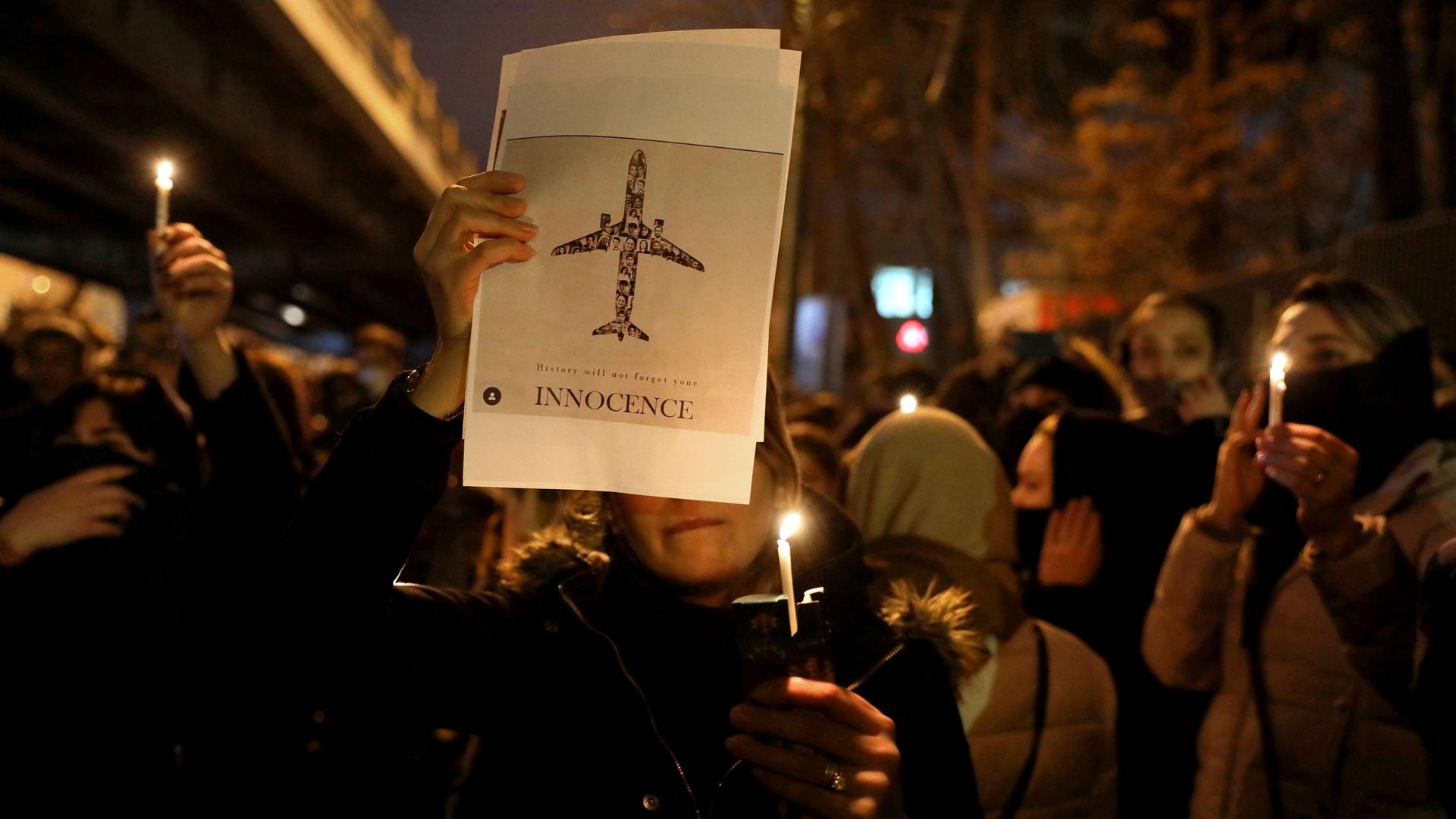 A crowd of people are shown at night holding candles with one person holding a poster of an aircraft with the word "Innocent" printed on it.