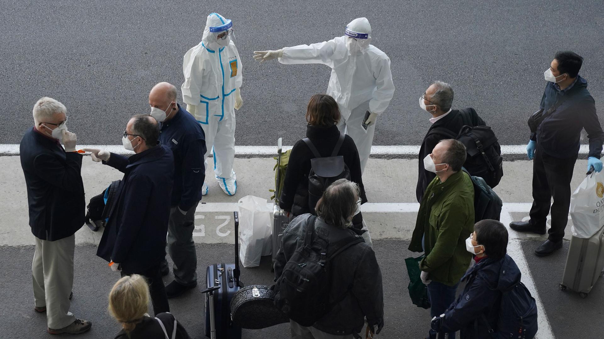 A small crowd of people are shown standing on a sidewalk with two people wearing white medical protective clothing and face masks.
