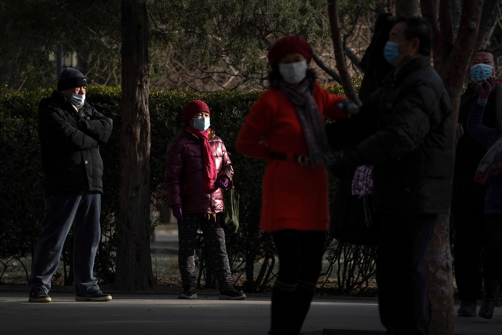 Several people are shown standing in a park and wearing face masks and winter jackes=ts.