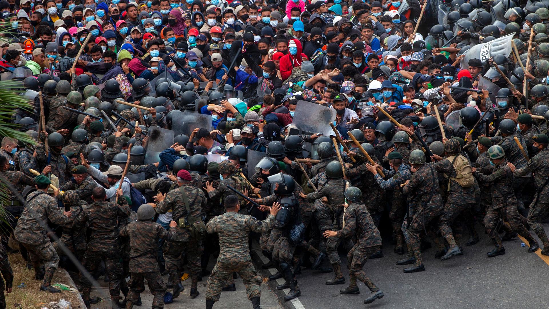 A large crowd of people are shown in conflict with security officials wearing military fatigues and helmets in a road.