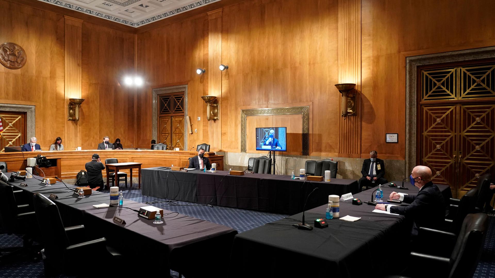 Homeland Security Secretary nominee Alejandro Mayorkas is shown seated at a long, cloth covered table in a largely empty room with tall, wooden walls.
