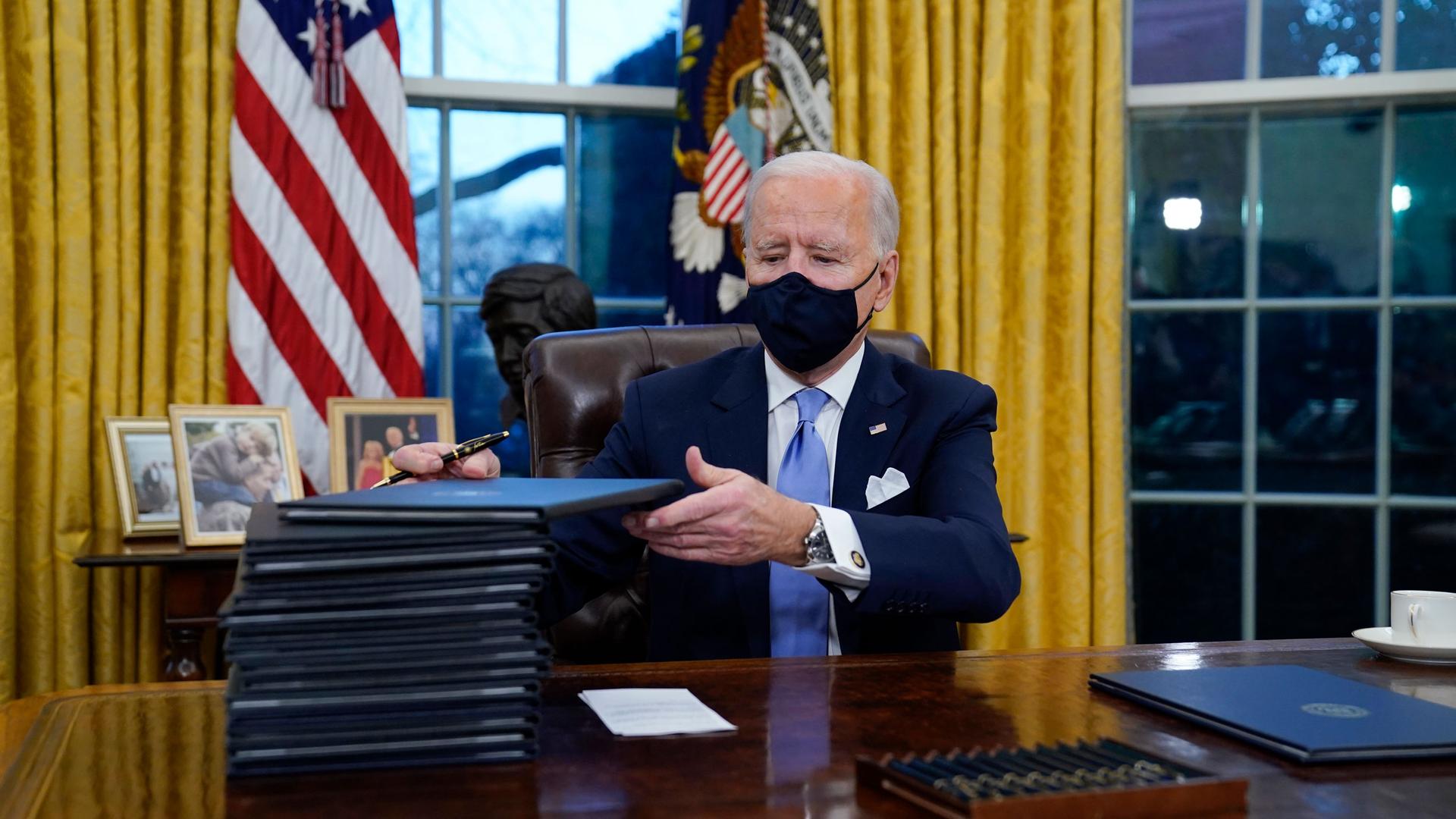 US President Joe Biden is shown seated at a wooden desk with a tall stack of embossed folders next to him.