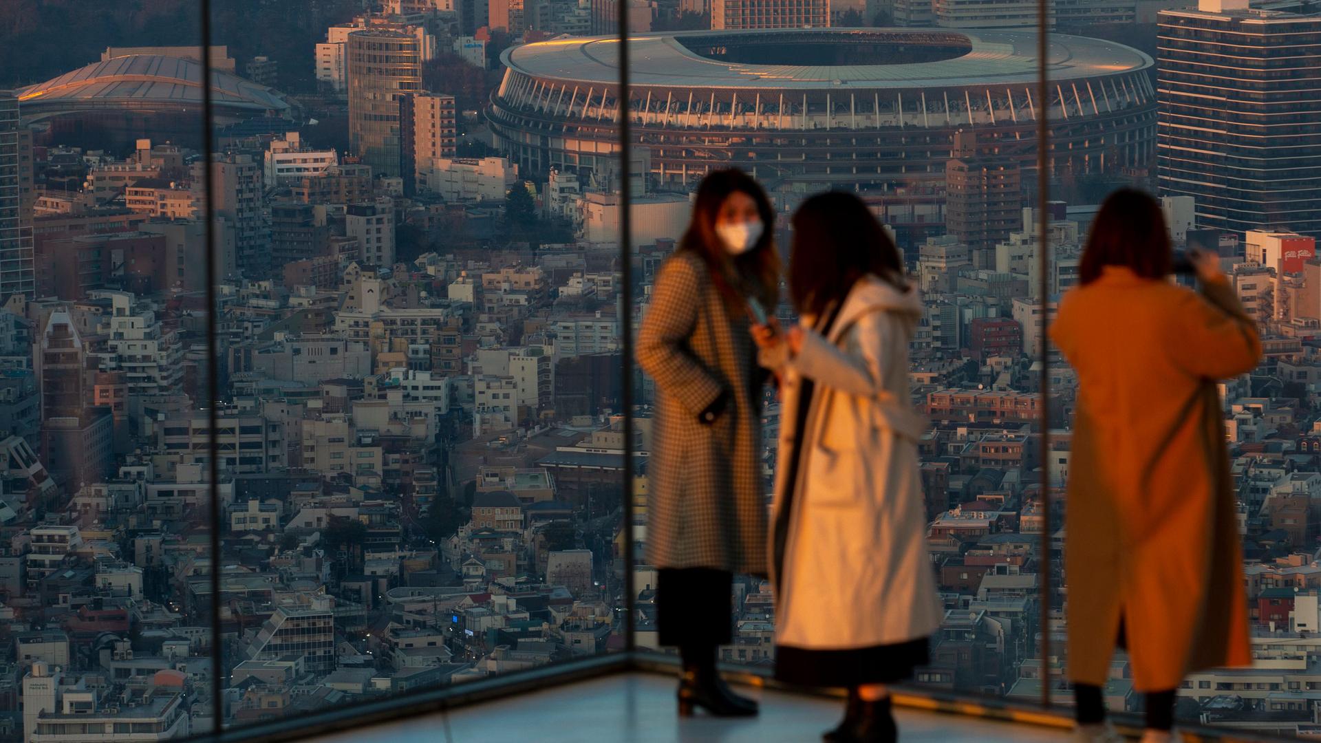 Japan National Stadium is shown in the distance from the vantage of a glass observatory where three women are standing.