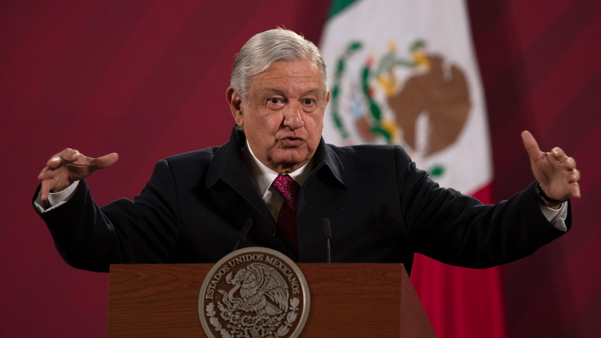 Mexican President Andrés Manuel López Obrador is shown speaking while standing behind a podium with his arms outstretched.