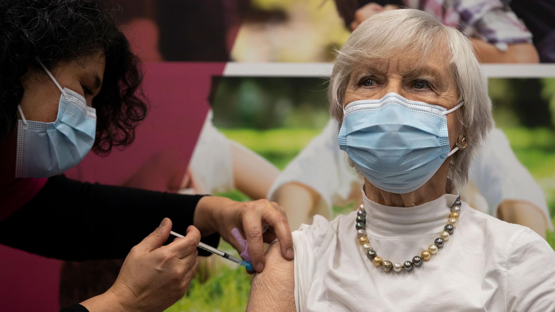 A woman is show wearing a white blouse with a pearl neclace and face mask while receiving a vaccine shot in her shoulder.
