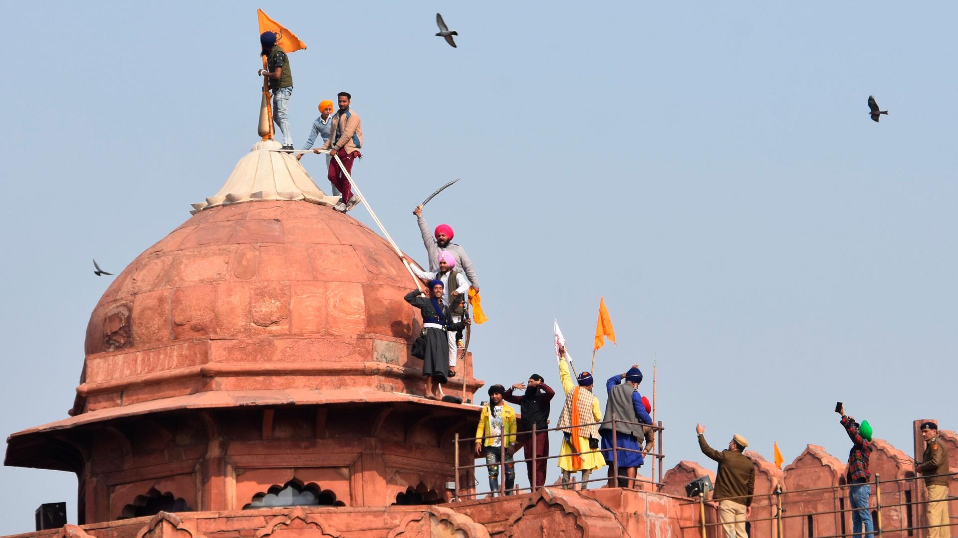 A group of men are shown on top of the redish-orange colored Red Fort monument with one man putting up an orange flag on top.