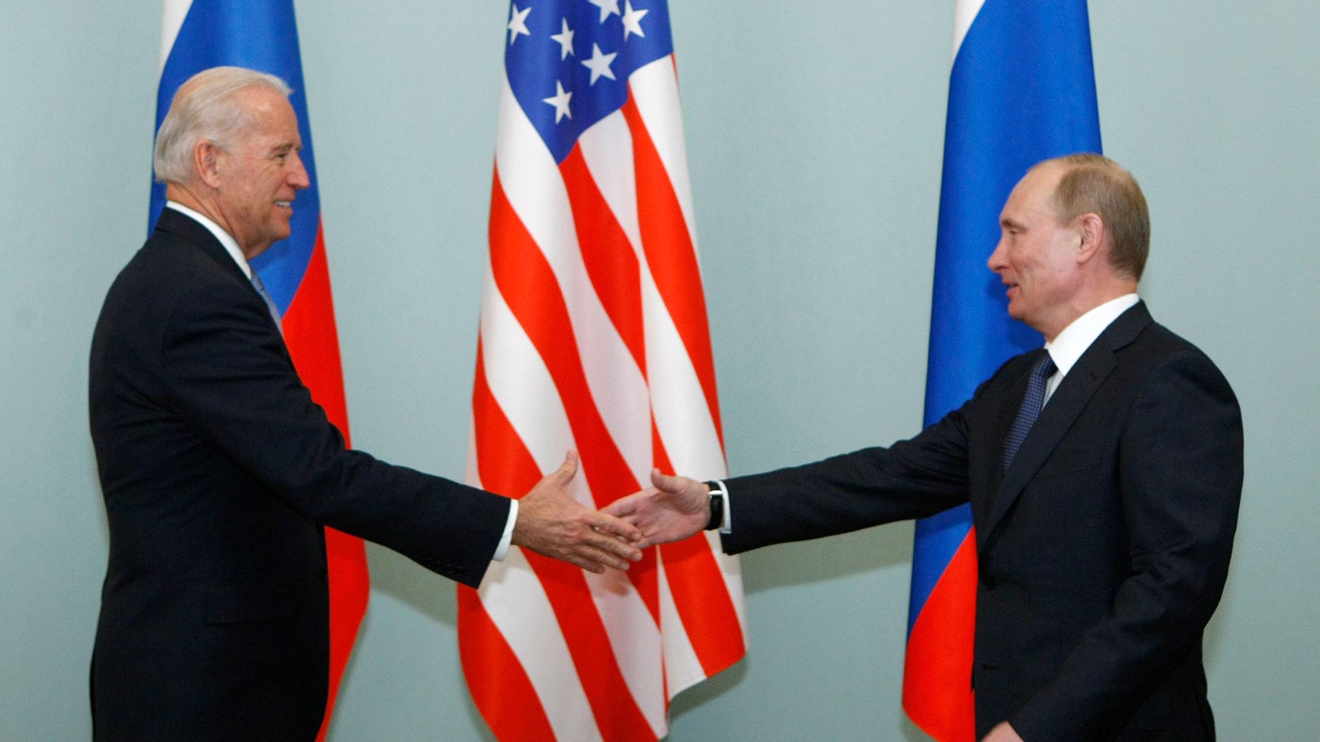 Joe Biden is shown with his hand outstretched shaking the hand of Vladimir Putin.