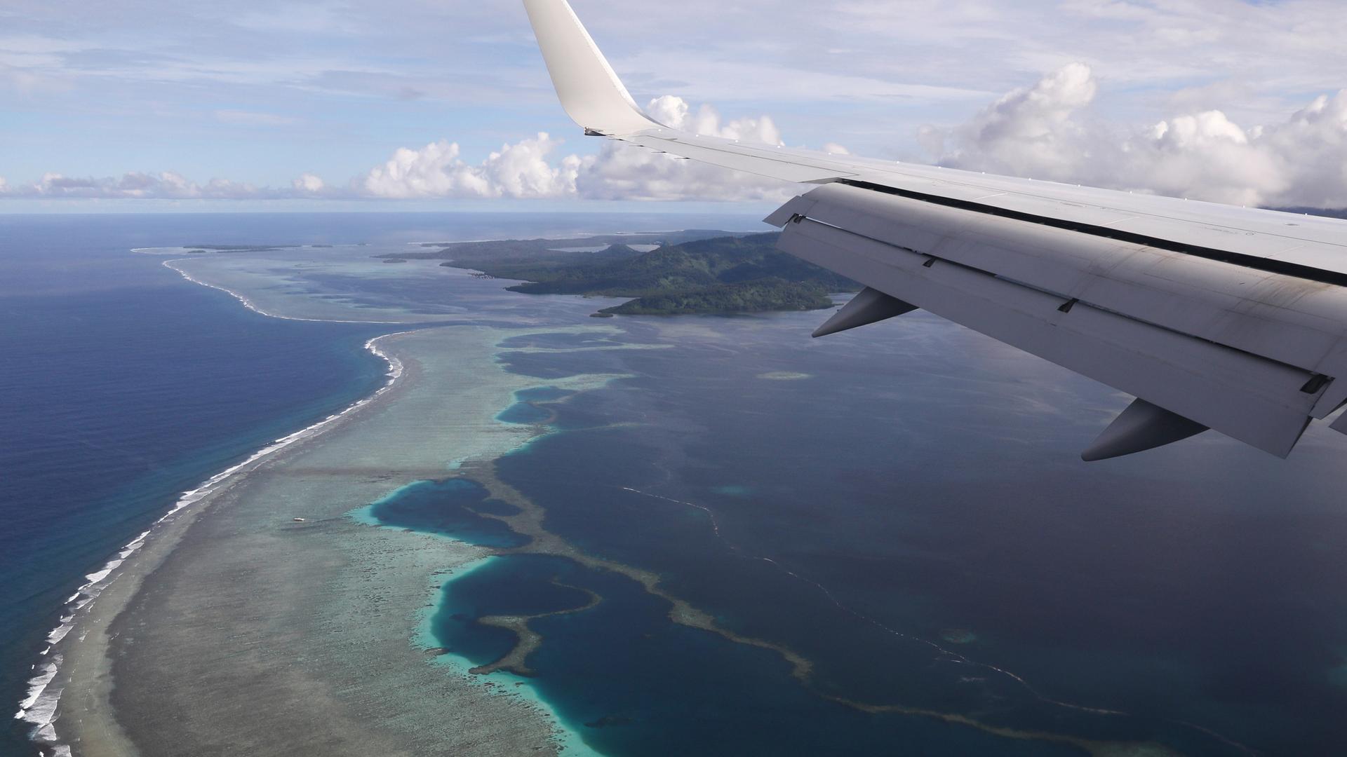 A plane flies over blue ocean waters and small island strips. 