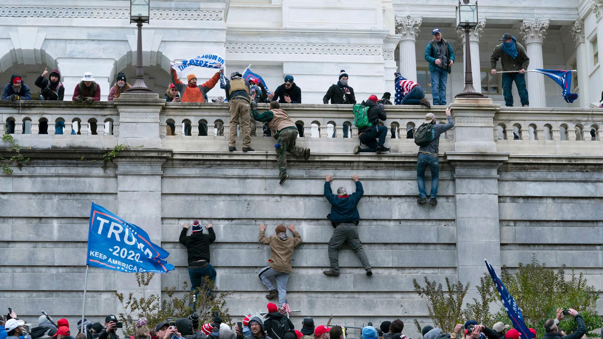 A large group of rioting supporters of former President Donald Trump are show carrying Trump 2020 flags and climbing the stone wall of the US Capitol.