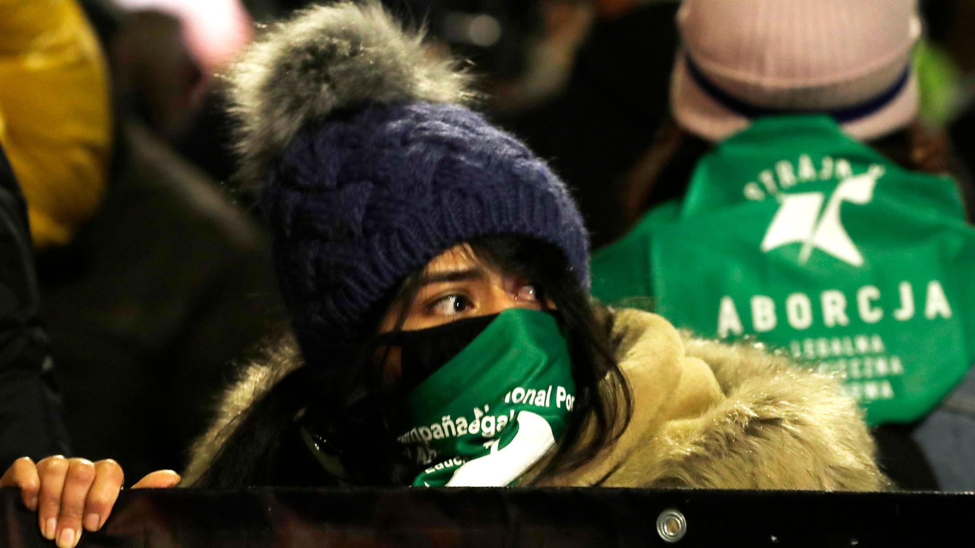 A crowd of people are shown with a woman in the center holding a banner and wearing a winter hat and green scarf with writing on it.