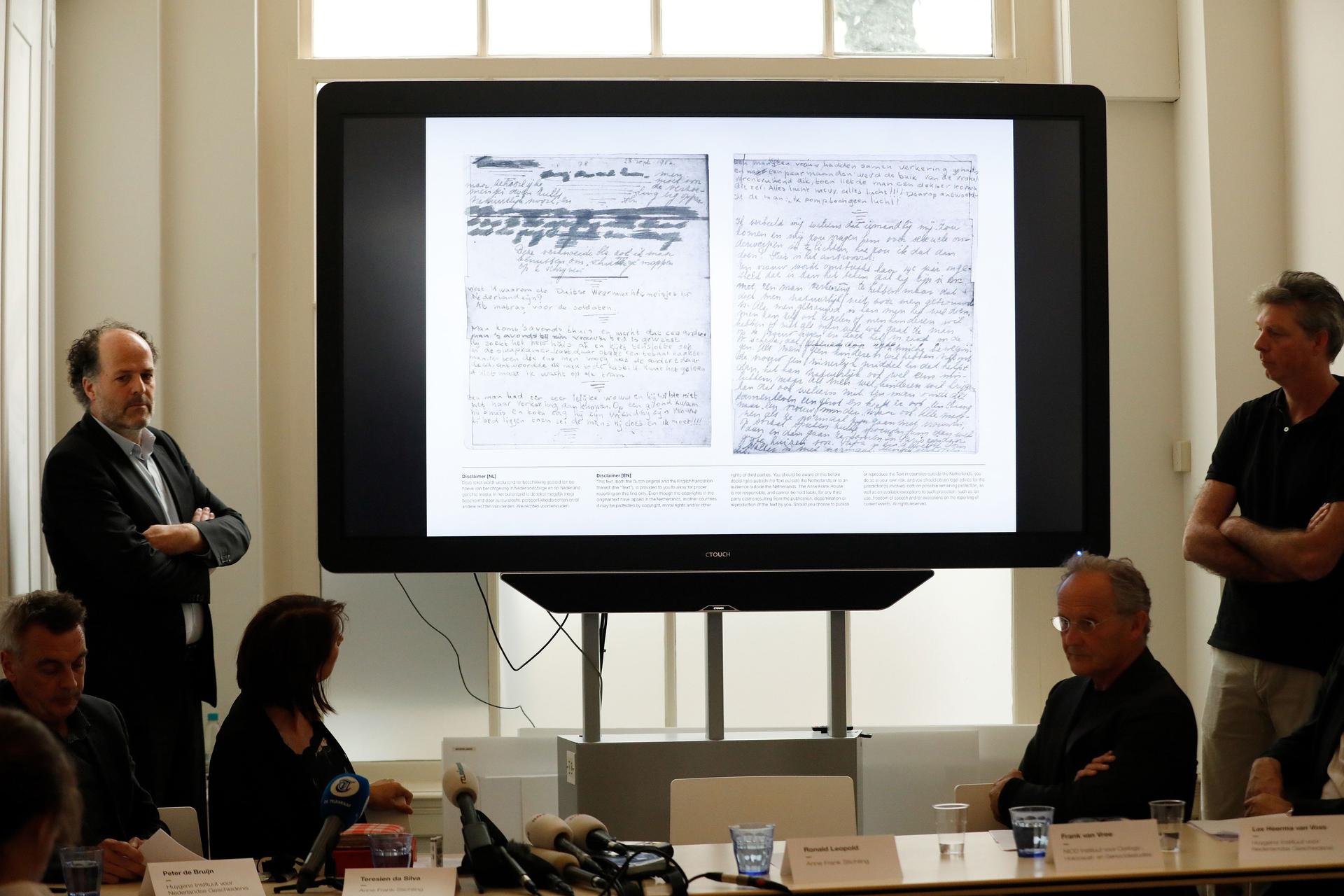 Two men stand on both sides of a projector screen, displaying pages of Anne Frank's handwriting. 