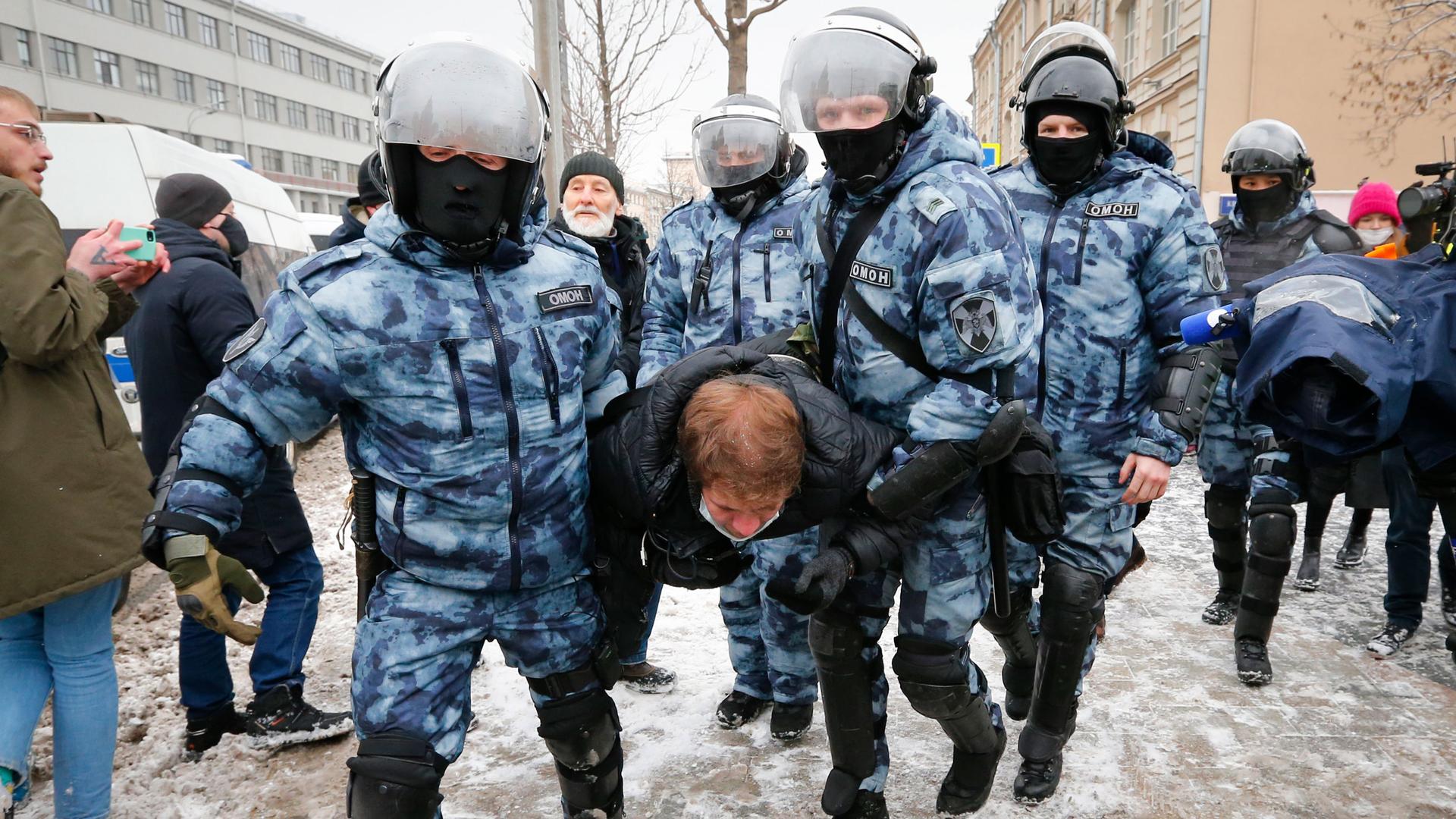 Several police officers are shown wearing winter camoflage and helmets while carrying a man by his arms and legs.