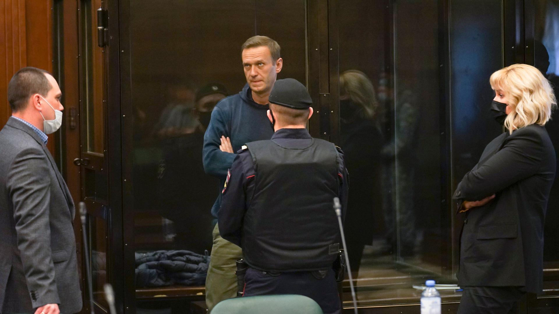 Alexei Navalny is shown standing inside of a clear glass cage in a courtroom with three people on the outside looking in on him.