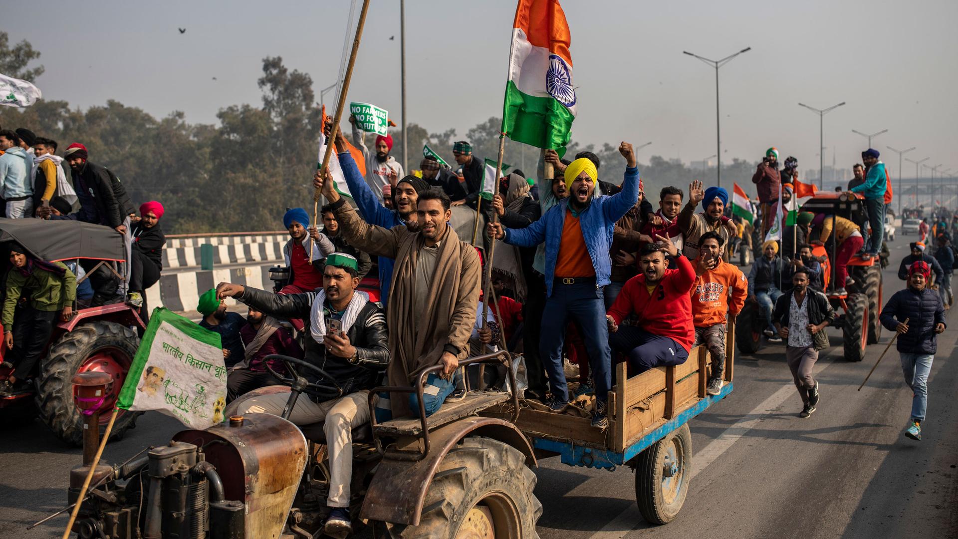 Protesters ride farm tractors in the road while carrying flags.
