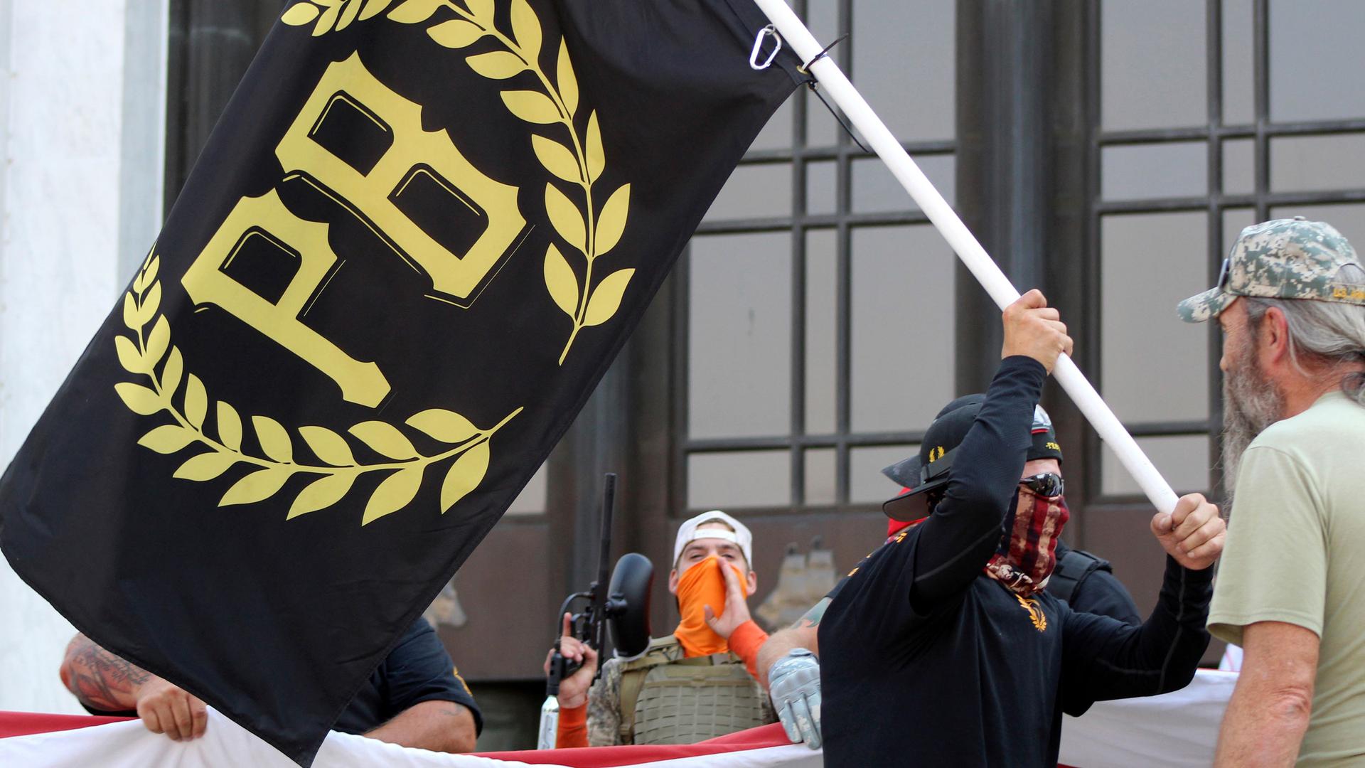 A man is shown wearing a face covering and holding a black flag with gold lettering with the letters P and B on it.