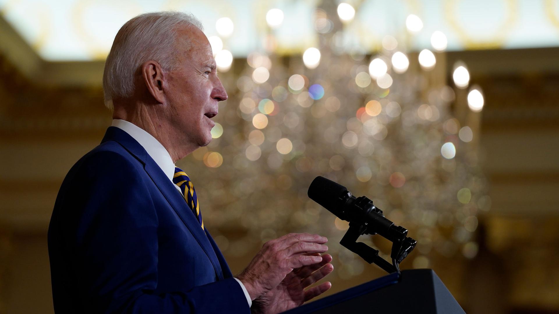 President Joe Biden is shown in profile speaking at a podium with microphones and wearing a blue suit.