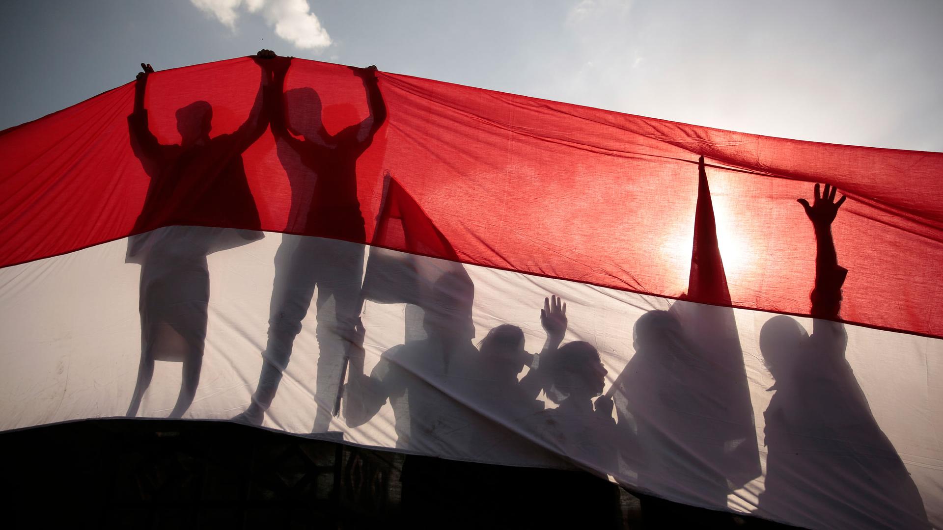 The dark silhouetted of several people are shown with their hands in the air holding up the red, white and black-stripped Yemen flag.